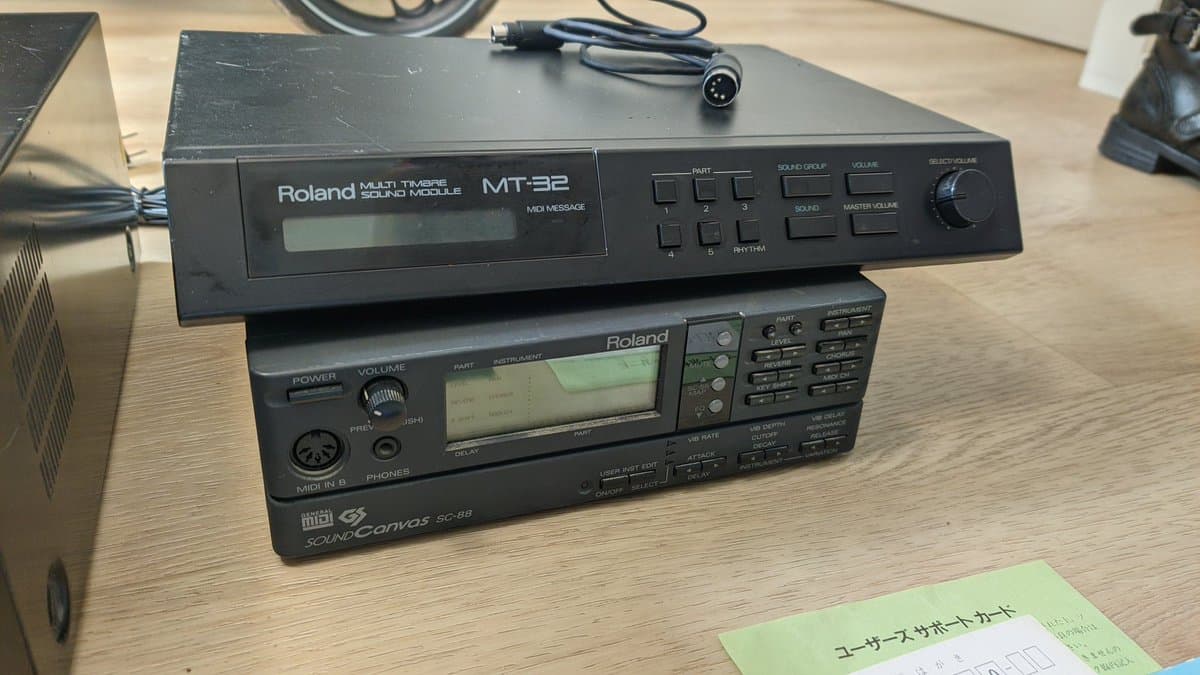 Roland MT-32 and SC-88 Sound Canvas MIDI modules stacked on wooden desk with cables and Japanese user manual visible.