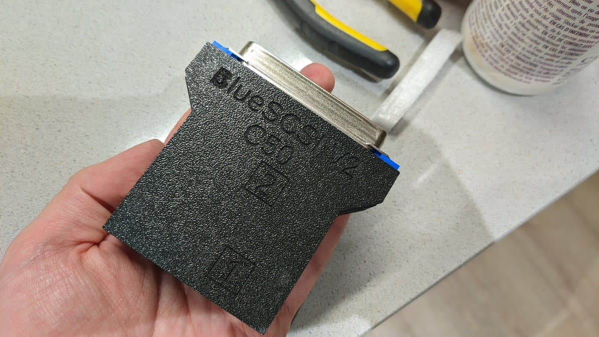 Hand holding assembled BlueSCSI v2 board with thin plastic shell, showing textured surface and metal connector.