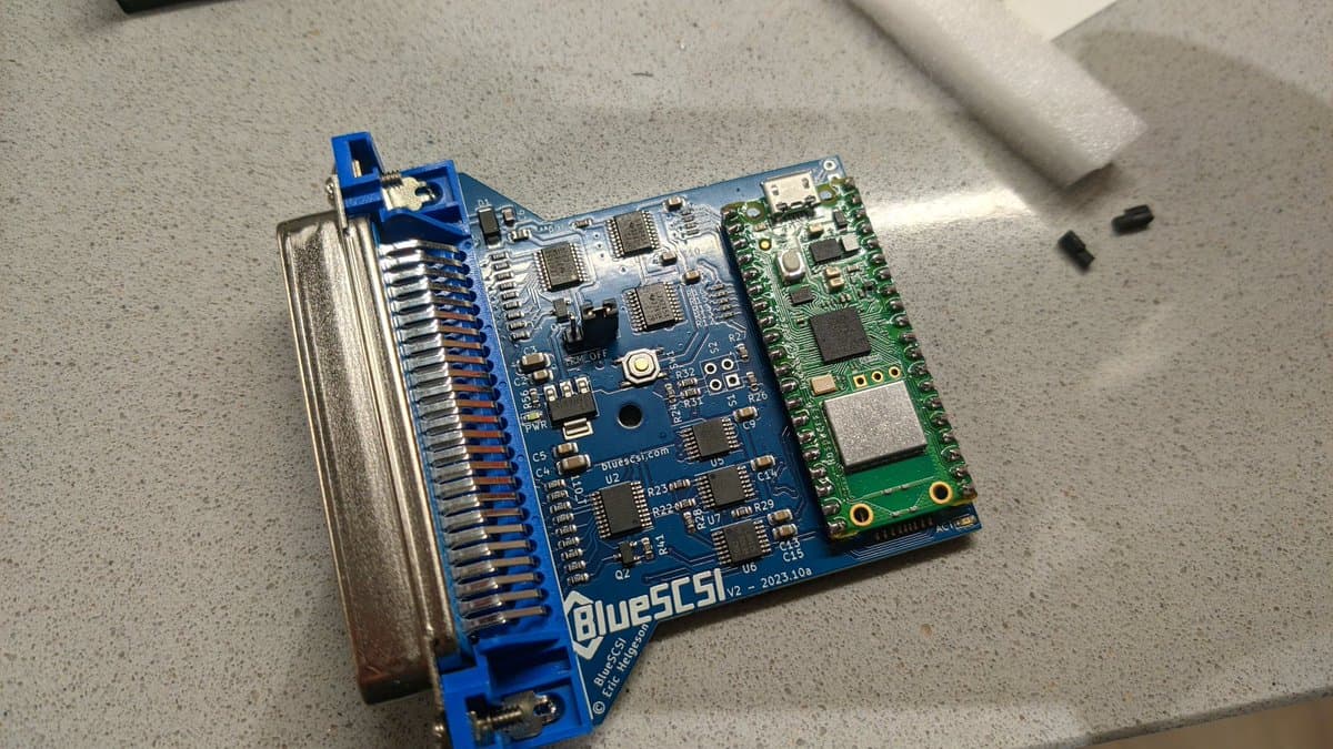 BlueSCSI v2 board assembly with blue SCSI connector, microcontroller, and green daughter board module on concrete surface.