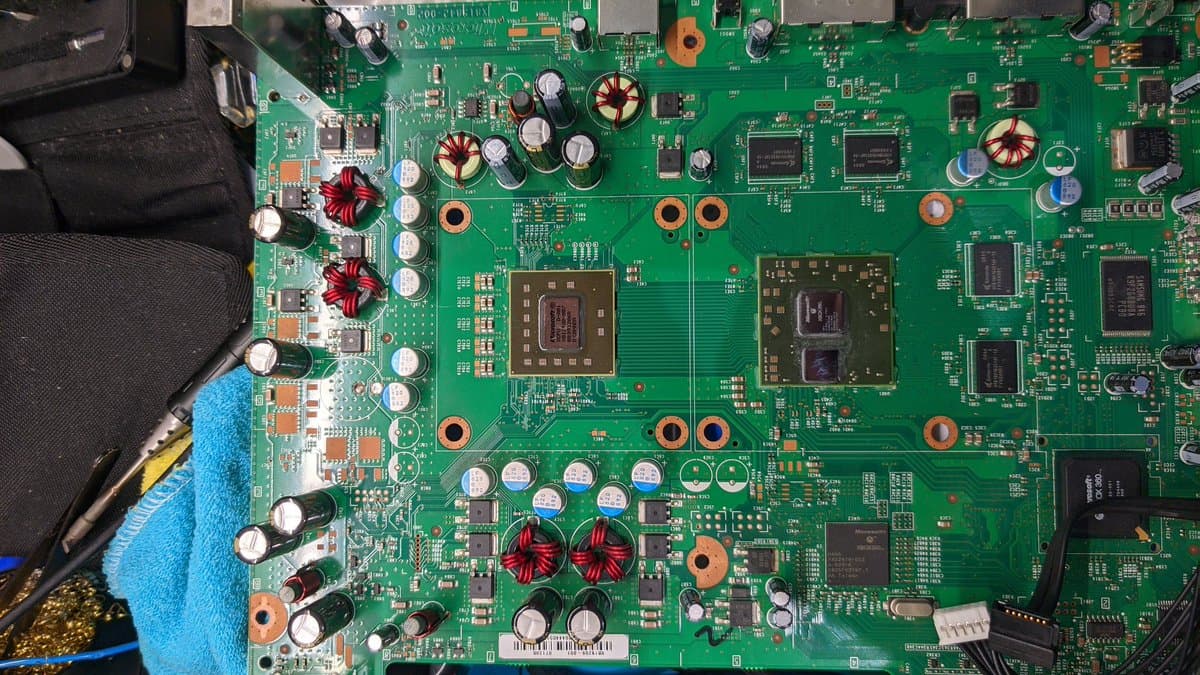 Xbox 360 mainboard during repair, showing dual GPU chips, power inductors, capacitors, and laser assembly area being serviced with cleaning tools.