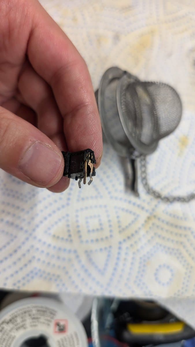 Corroded Sega Wondermega power jack held in fingers, showing oxidation damage; connector and cable visible blurred in background.