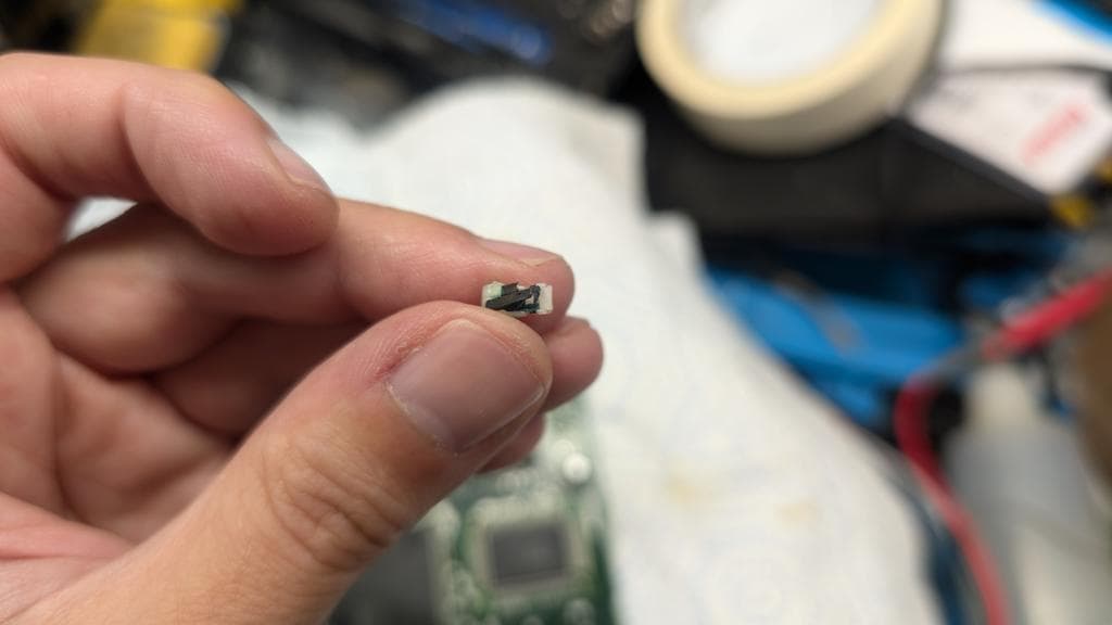 Corroded cartridge detection button held in tweezers over workbench with tape and repair supplies visible.