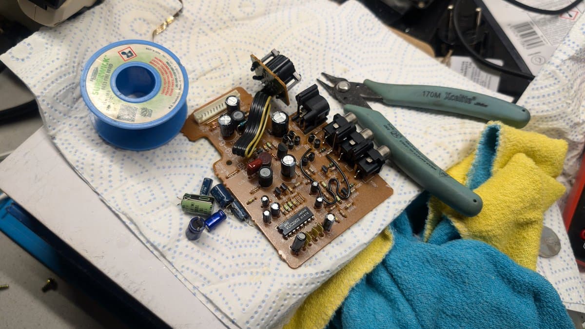 Wondermega AV board partially recapped with new capacitors, reflowed solder joints, soldering iron, spool of solder, and yellow-blue cloth on work surface.