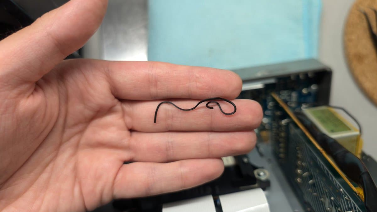 Hand holding a worn rubber belt shaped like a coiled snake against a workbench with electronic equipment in the background.