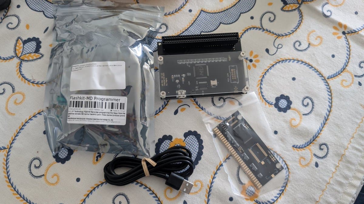 Flashkit-MD Programmer kit laid out on patterned fabric: programmer unit in black case, USB cable, and RAM cartridge in protective bags.