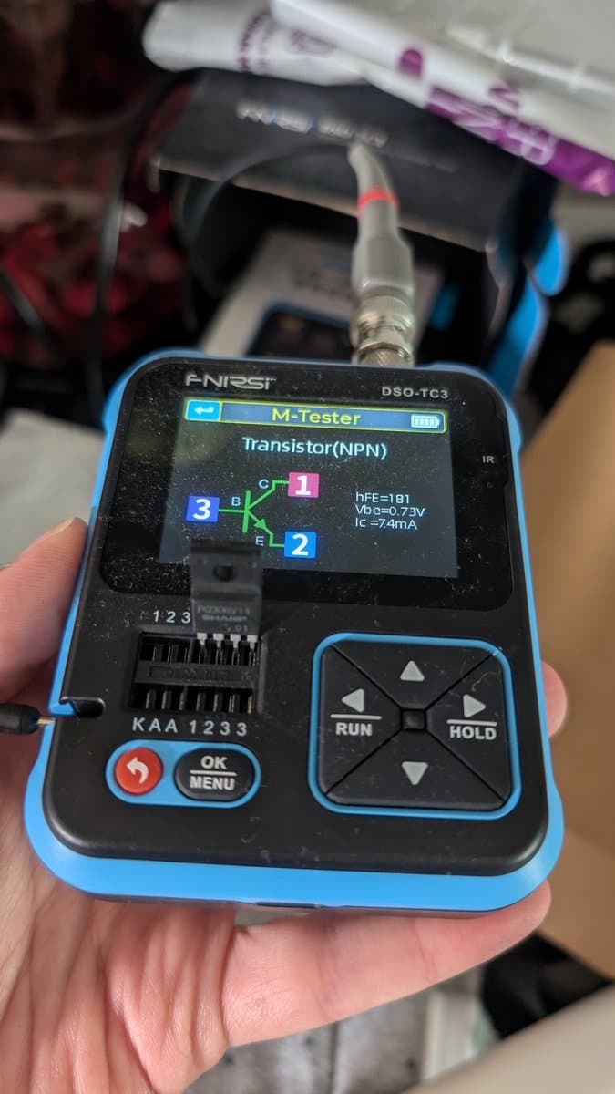 ANRST M-Tester displaying NPN transistor test results (hFE=181, VBE=0.73V, IC=74mA) with probe connected to component legs during Wondermega repair diagnostics.