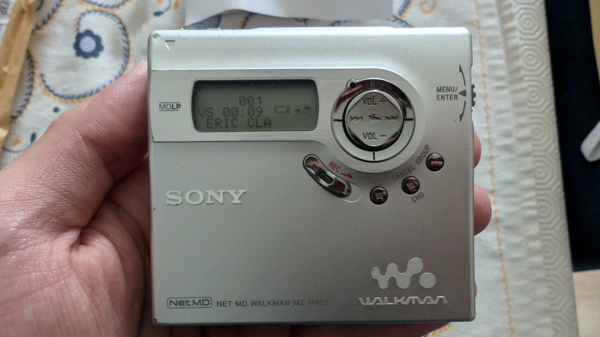 Sony Net MD Walkman MZ-N920 held in hand, displaying LCD screen showing time and "ERIC CLA" track info, silver casing visible with volume controls.