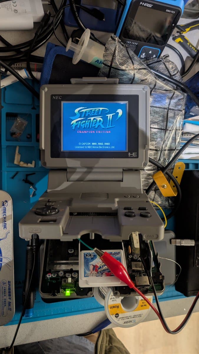 NEC PC Engine LT with open chassis, displaying Street Fighter II on screen, bench PSU with red probe testing voltage to LCD panel during repair diagnostics.