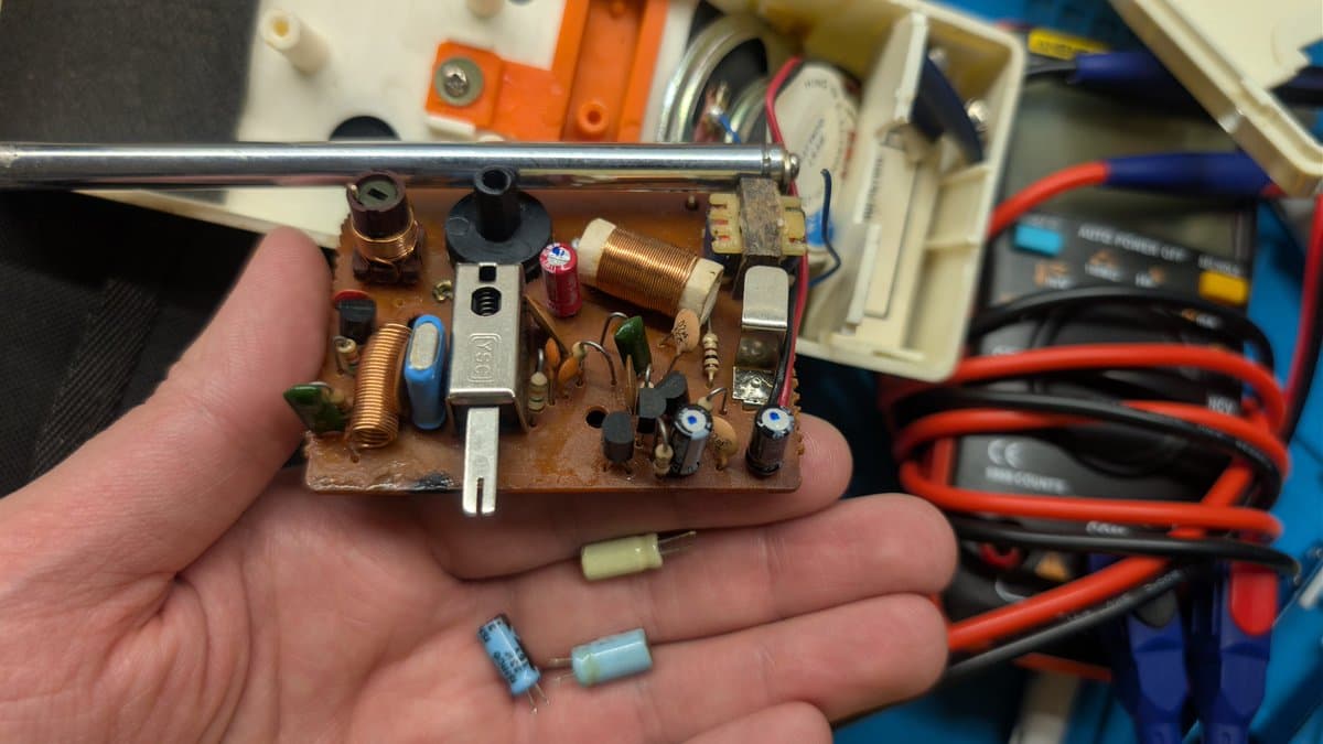 Disassembled walkie-talkie PCB held in hand showing recapped components, coil, capacitors, and corrosion cleaning work during restoration.