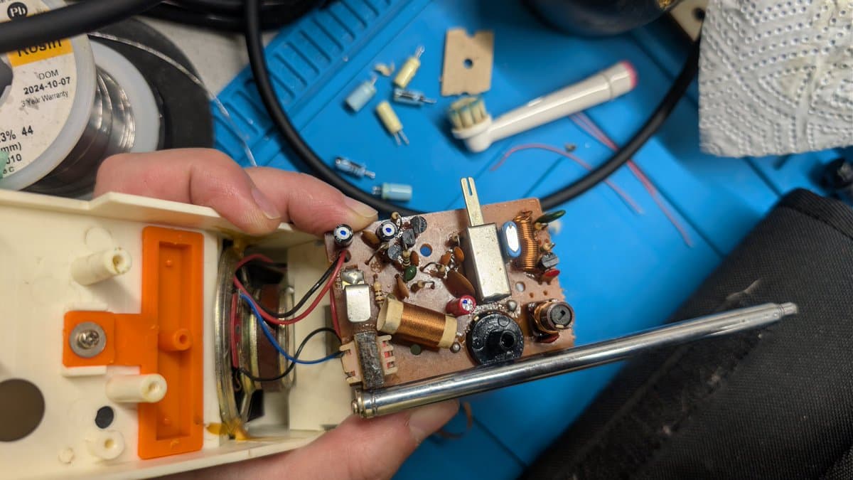 Disassembled walkie-talkie PCB held in hand showing capacitors, coils, variable resistor, and solder joints during refurbishment.