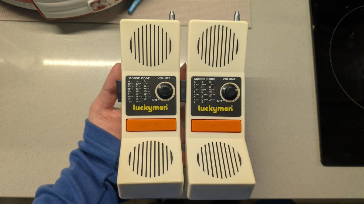 Two restored "Luckyman" walkie-talkies held in hand, showing cream plastic housing, speaker grilles, morse code charts, volume dials, and orange battery covers.