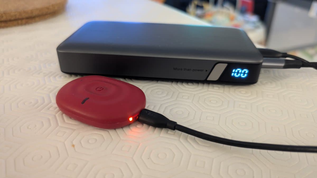 Red PowerDot EMS device with charging cable connected to black power bank showing 100% charge display on white patterned surface.