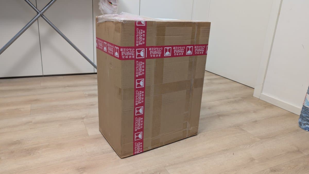 Unopened cardboard shipping box with red fragile tape and Japanese text, sitting on wooden floor next to white wall.