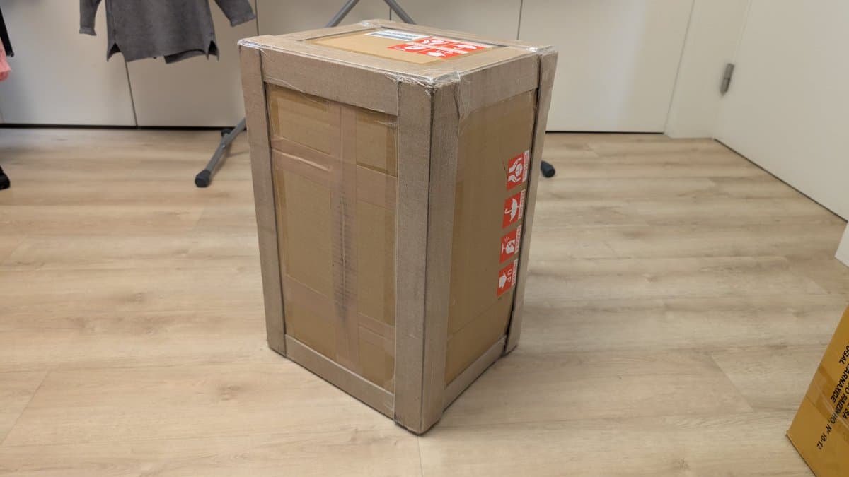Large cardboard shipping box sealed with tan packing tape on wooden floor, red shipping labels visible on side.