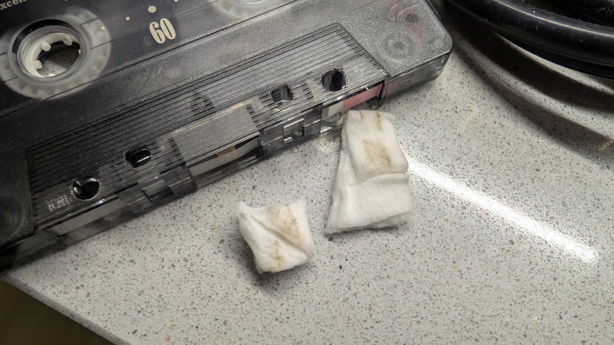 Cassette tapes being cleaned with white cloth and black cassette shell showing tape reels and label visible on workbench.