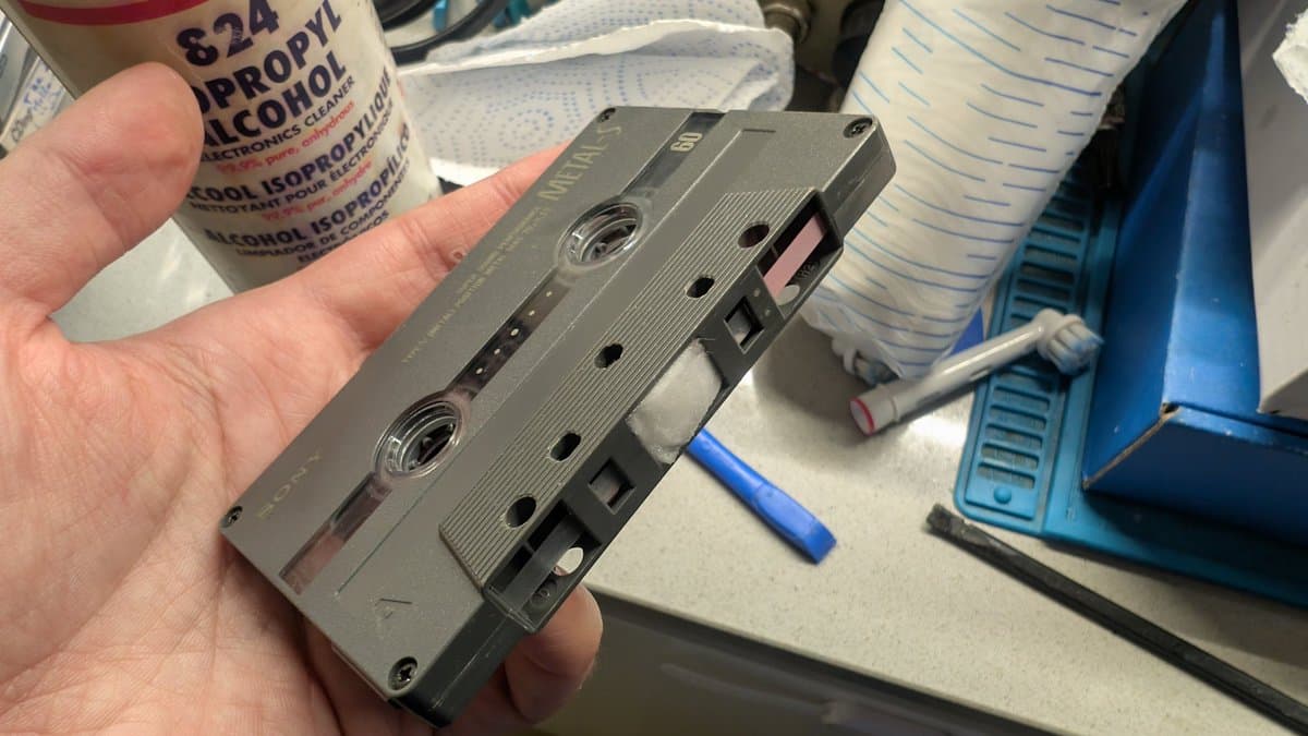 Hand holding gray cassette tape during cleaning, isopropyl alcohol and cleaning supplies visible on workbench.