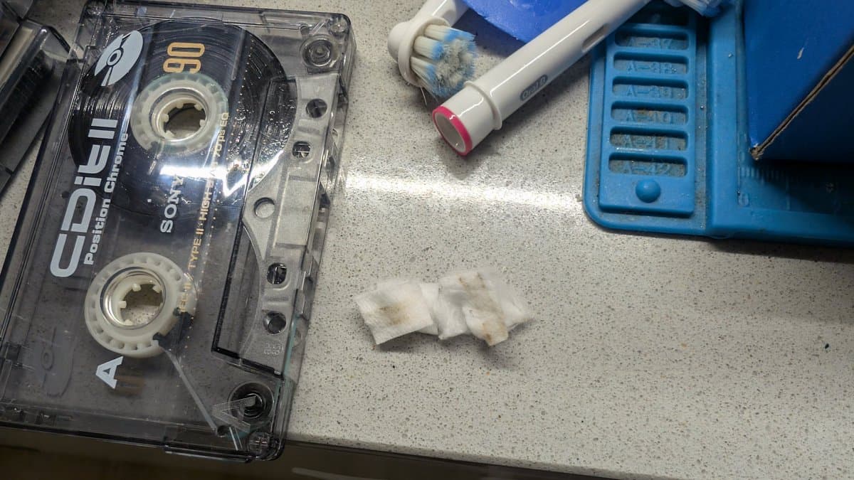Cassette tape maintenance setup: Akai Type II cassette with cleaning supplies, cotton swabs, and blue tape deck on workbench surface.
