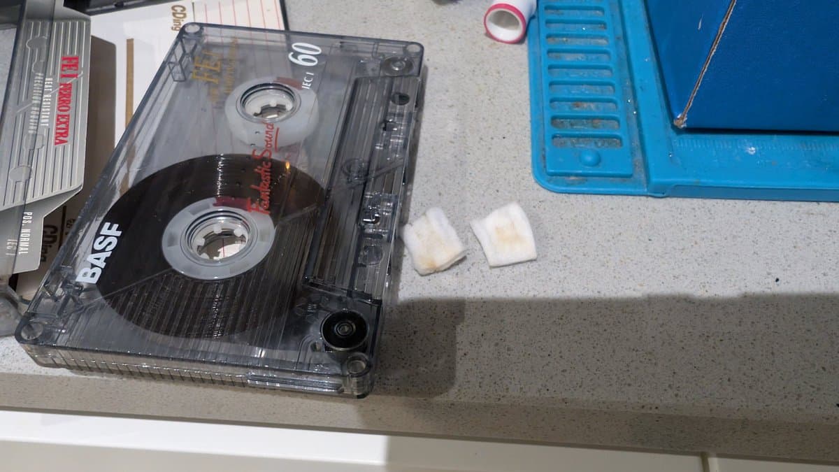 Cassette tape on work surface with cleaning supplies and cotton swabs nearby; blue storage container and documentation visible.