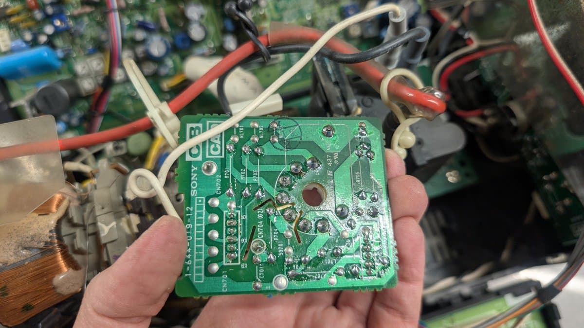 Hand holding Sony PVM-6041Q neck board showing dull solder joints and components, with other circuit boards blurred in background.