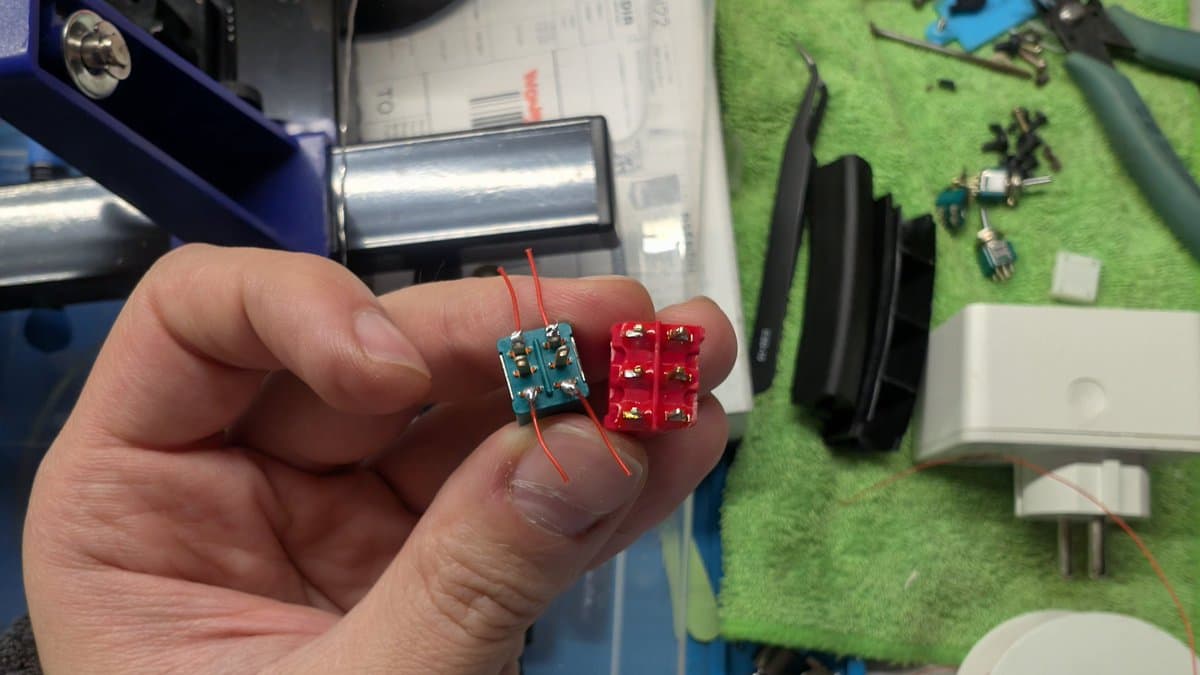 Hand holding blue and red RGB mod PCBs with wire leads soldered, workbench with components on green mat in background.