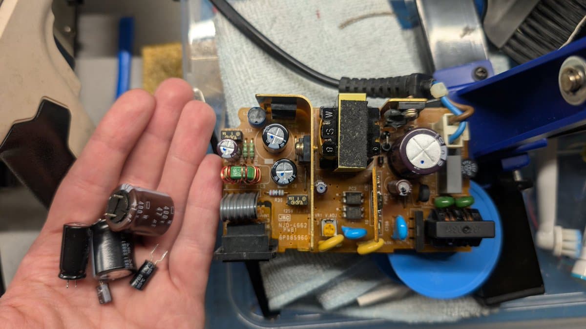 Technician's hand holding removed capacitors next to N64 power supply board during recap service, showing desoldered components.
