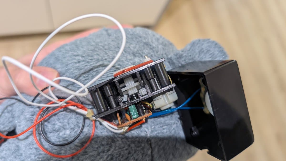 Close-up of Totoro toy mechanism showing exposed gears, motor, capacitors, and red/white wires on gray fabric backing during repair.