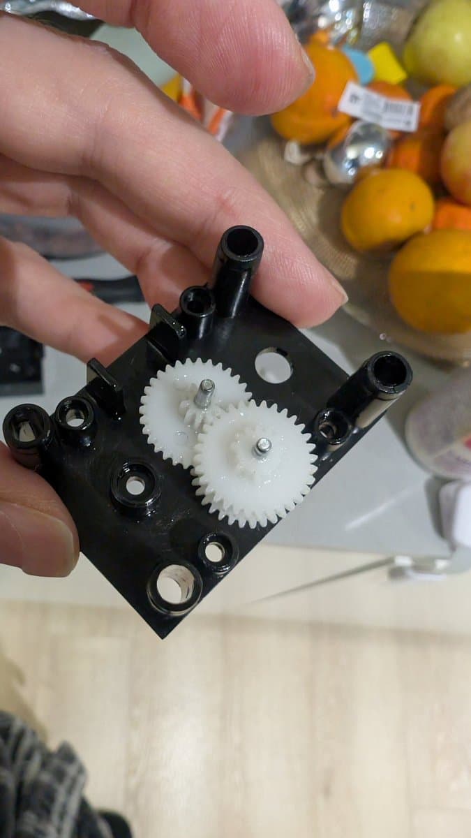 Close-up of black plastic gear assembly with two white gears meshed together, mounted on black housing with metal posts and mounting holes; citrus fruits blurred in background.