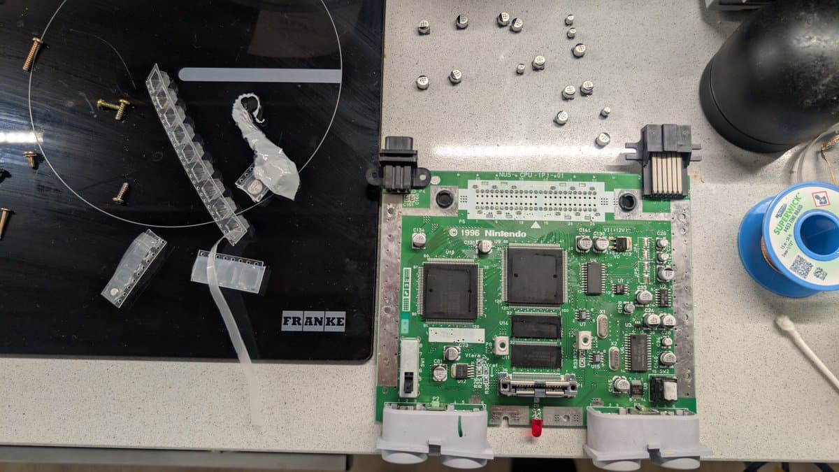 Nintendo 64 motherboard with replaced capacitors, original PCB with brass hardware visible, soldering iron and solder nearby on work surface.