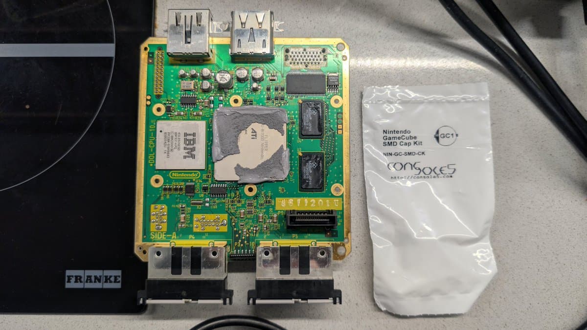 Nintendo GameCube mainboard (side A) with exposed CPU and RAM chips, two HDMI ports, and SMD cap kit package during recap restoration project.