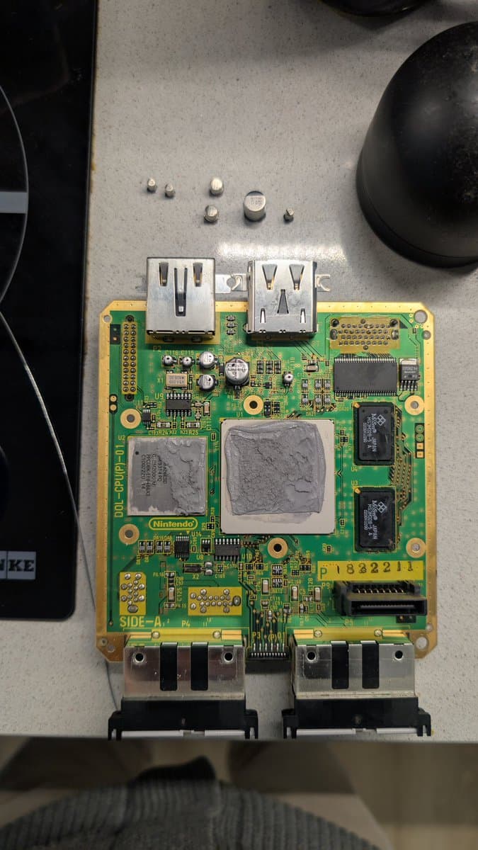 Nintendo GameCube DOL-001 motherboard with recapped optical drive, two USB ports visible, dust on heatsinks, loose screws above board.