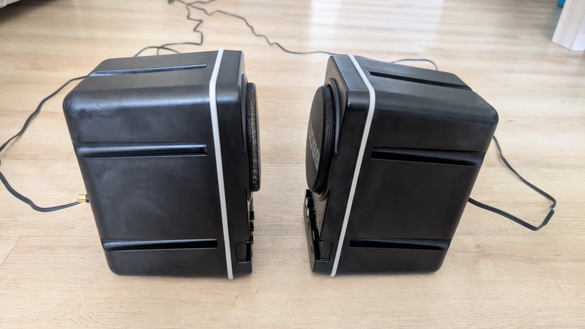 Pair of refurbished BOSS MA-12AV speakers with white trim on wooden floor, showing cleaned black plastic enclosures and speaker grilles after maintenance.