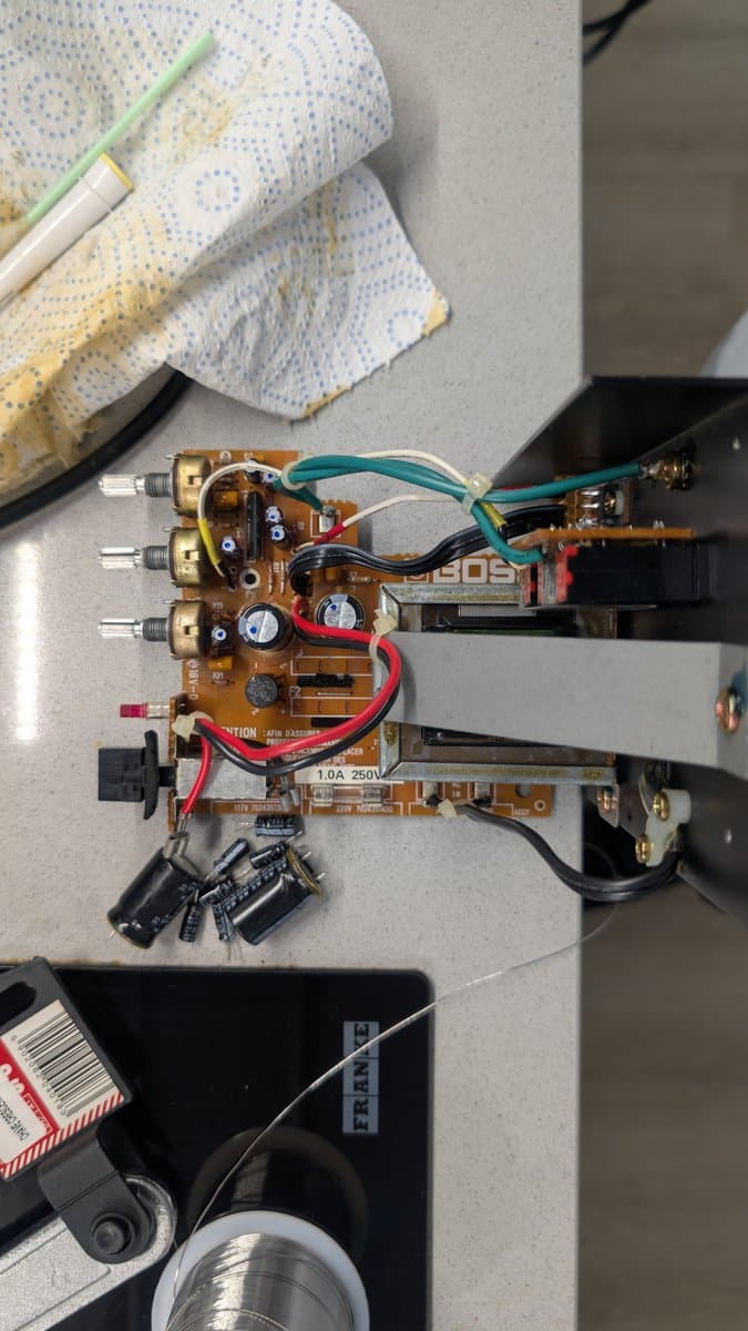 Recapped BOSS MA-12AV amplifier board with exposed circuit components, capacitors, and wiring during maintenance; paper towels and brush visible above.