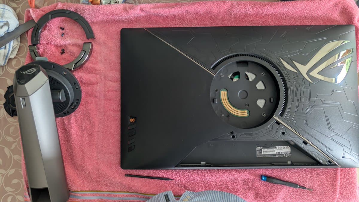 Asus PG27UQ monitor bottom panel removed showing internal fan assembly with gold connectors and black shroud during refurbishment, headphones and screwdriver visible.