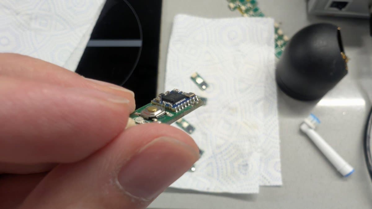 Finger holding a tiny PCB with soldered MCU chip, white schematic papers and black rework station visible in background.