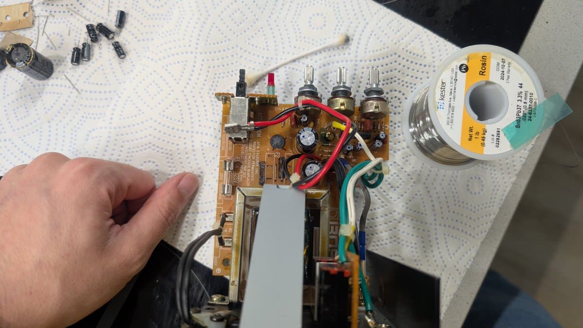 Close-up of BOSS MA-12AV amplifier board undergoing capacitor replacement and solder reflow, with solder spool and tools visible on workbench.