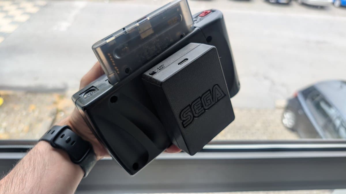 Hand holding Sega Nomad with aftermarket battery pack attached, showing restored portable console in daylight near window.