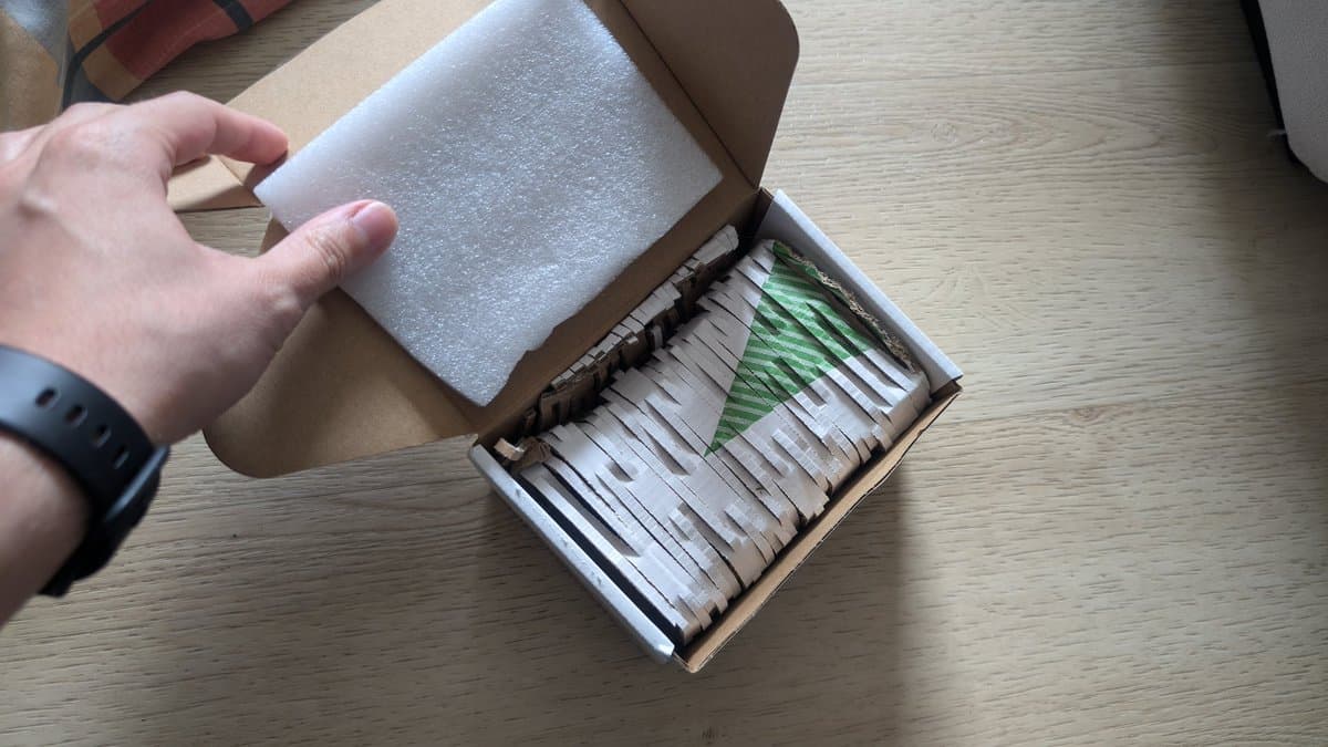 Hand opening box containing new aftermarket Nomad battery pack with foam padding and green branded label visible inside.