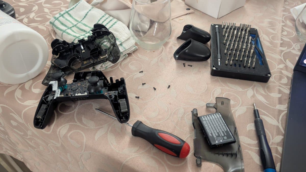 Disassembled Nintendo Switch Pro controller with circuit board exposed, screwdriver, bit set, and cleaning supplies on work surface.