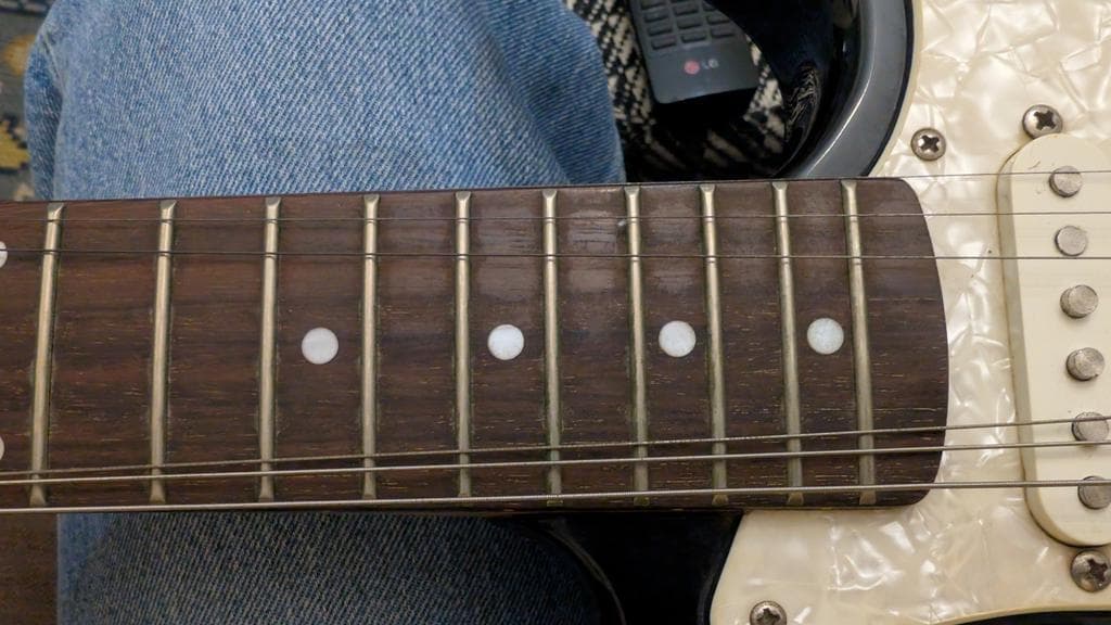 Close-up of electric guitar fretboard showing cleaned rosewood with inlay dots and nickel frets, cream pickguard visible.