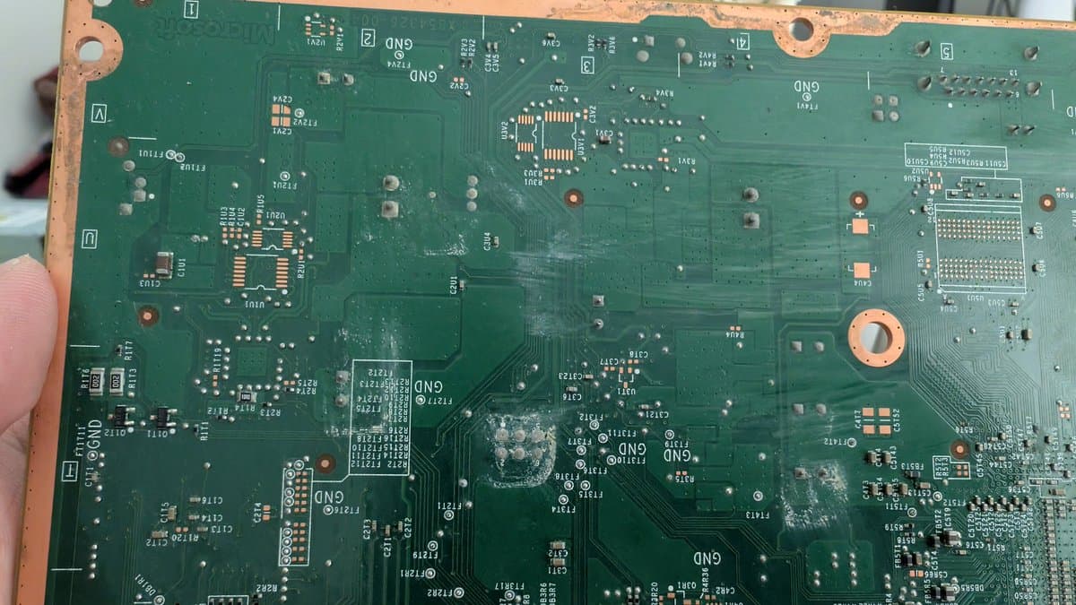 Xbox 360 E motherboard showing liquid damage residue and oxidation on components, with visible corrosion around IC chips and solder joints requiring cleaning and repair.
