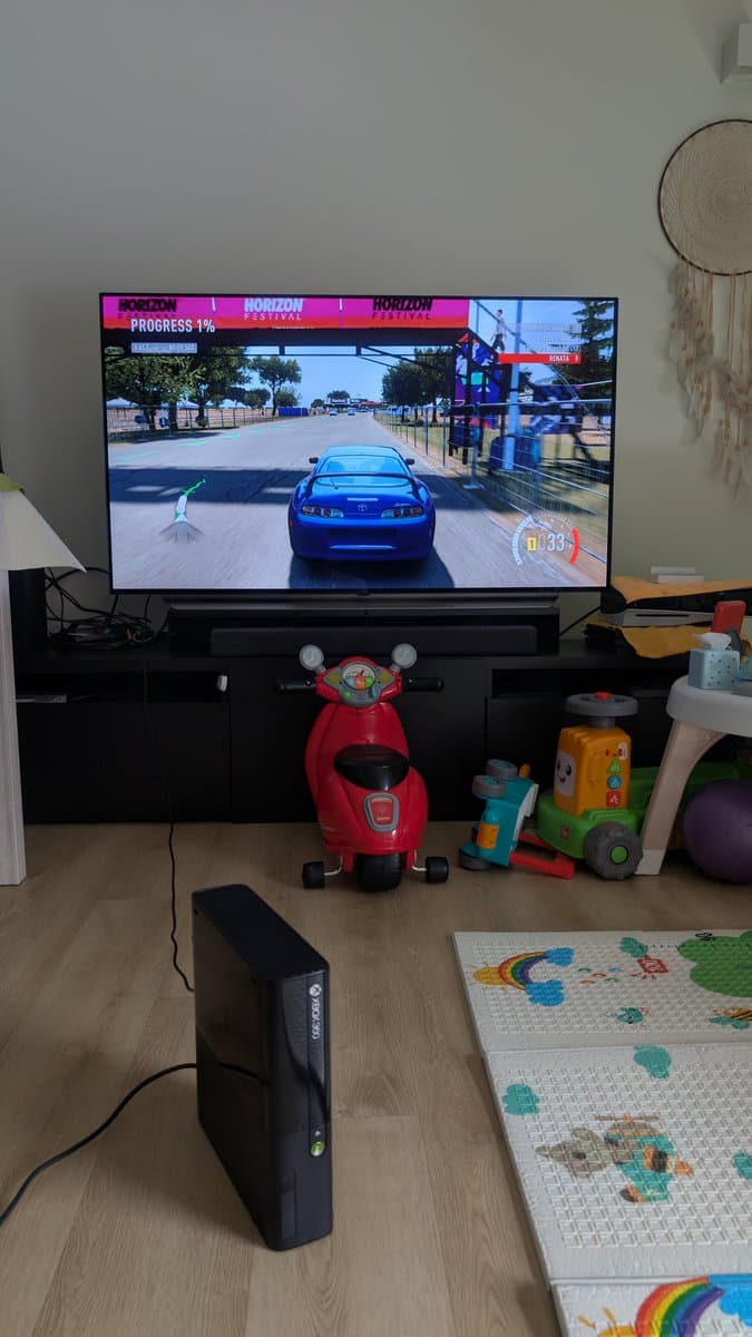 Xbox 360 E console displaying Forza Horizon game on TV, positioned on black entertainment stand with red toy scooter in front.