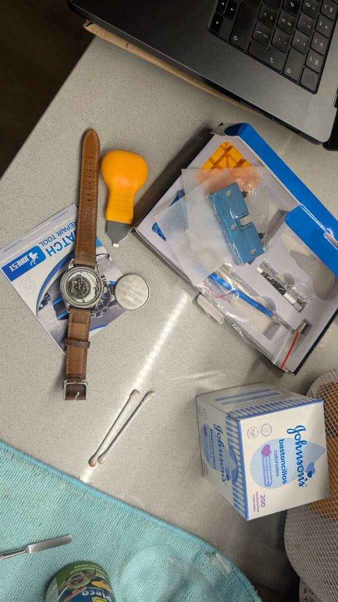 Watchmaking tools and parts laid out: watch with brown leather strap, yellow casing tool, magnifying glass, tweezers, blue model car kit, and batteries on desk workspace.