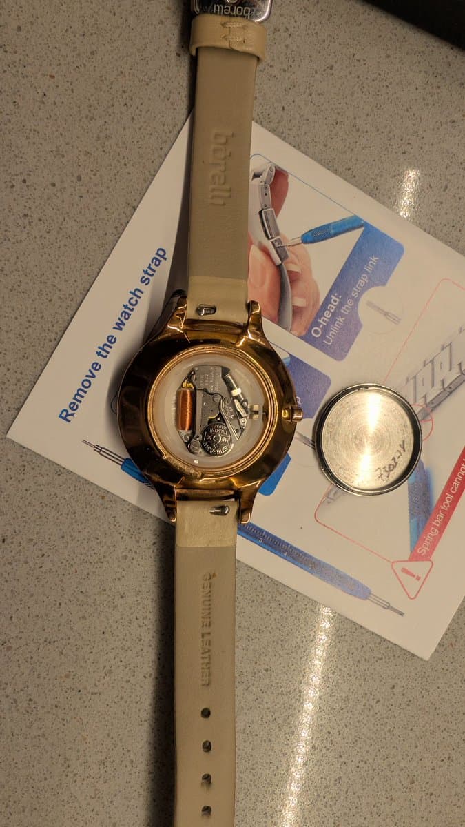 Disassembled watch with caseback removed, showing internal mechanism with balance wheel visible; repair tools and instruction manual laid beside it.