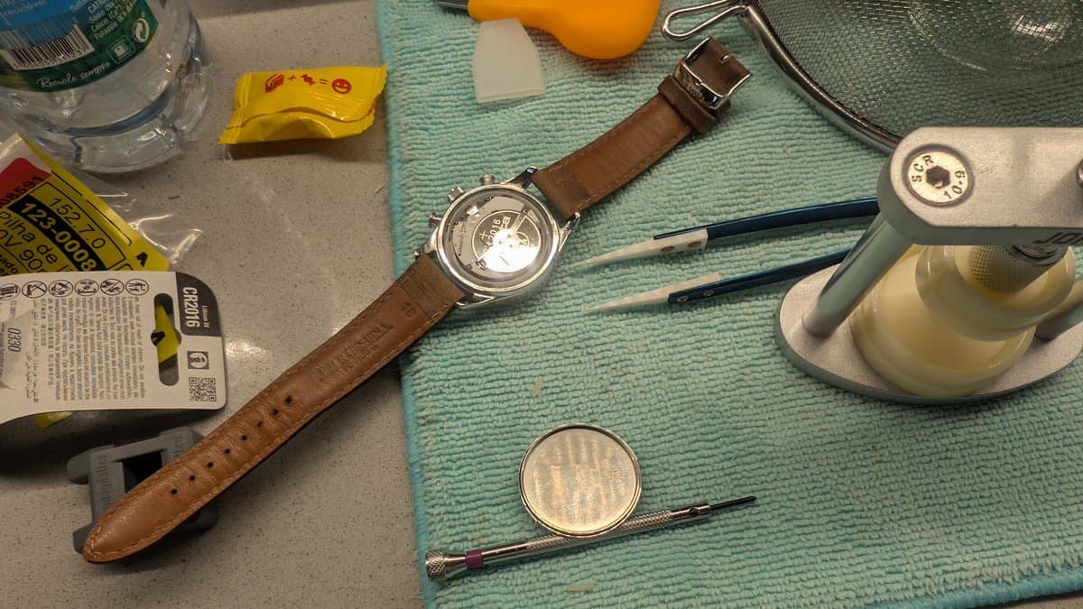 Watch battery replacement tools laid out: brown leather-strapped watch, case opener, tweezers, magnifying glass, and replacement batteries on turquoise work mat.