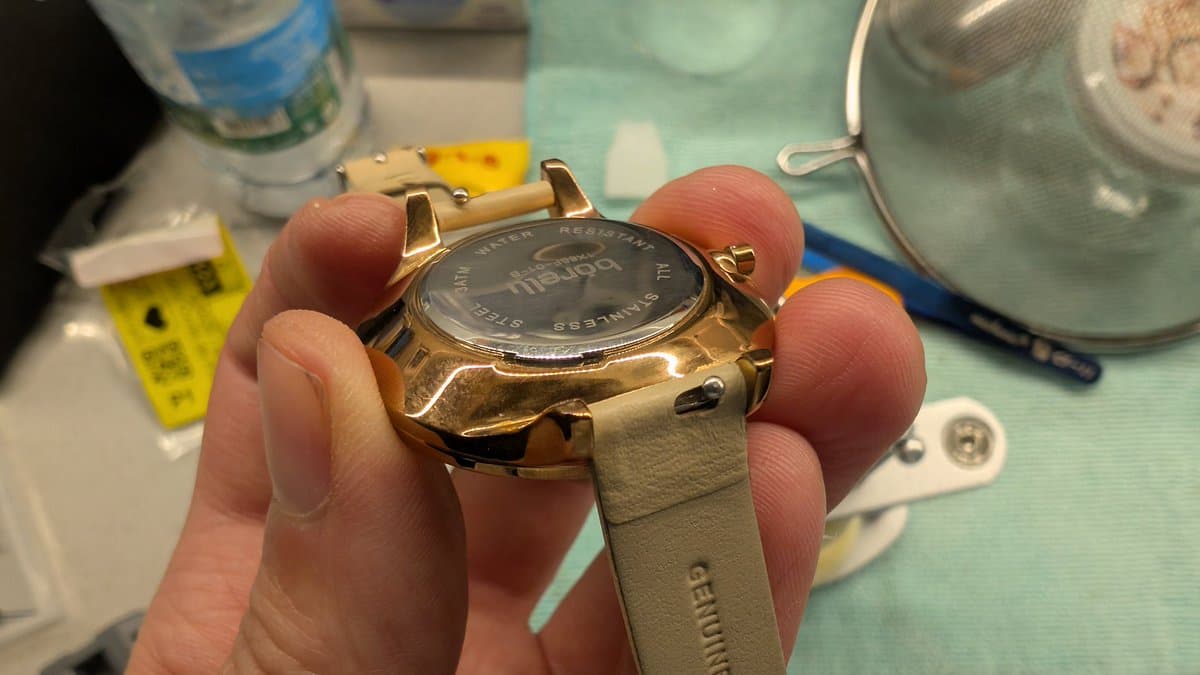 Gold-tone watch case held in hands during battery replacement, with case back removal tool and supplies visible on work surface.