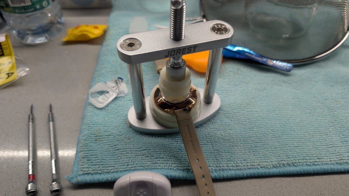Watchmaking tool setup with JDForest case press and adjustable wrench on blue mat, precision screwdrivers and calipers visible nearby.