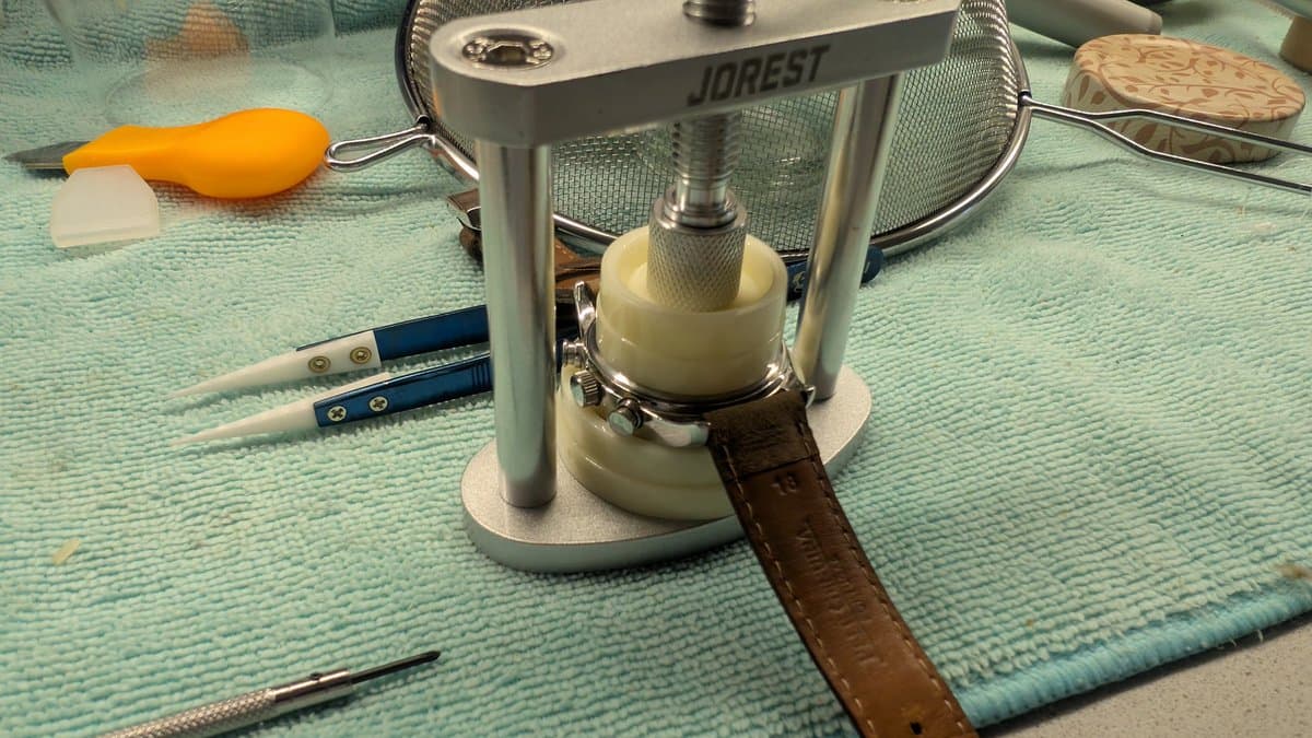 Jobrest watch case press tool with cream-colored platen, holding open watch case back; precision screwdrivers and orange battery case nearby on workmat.