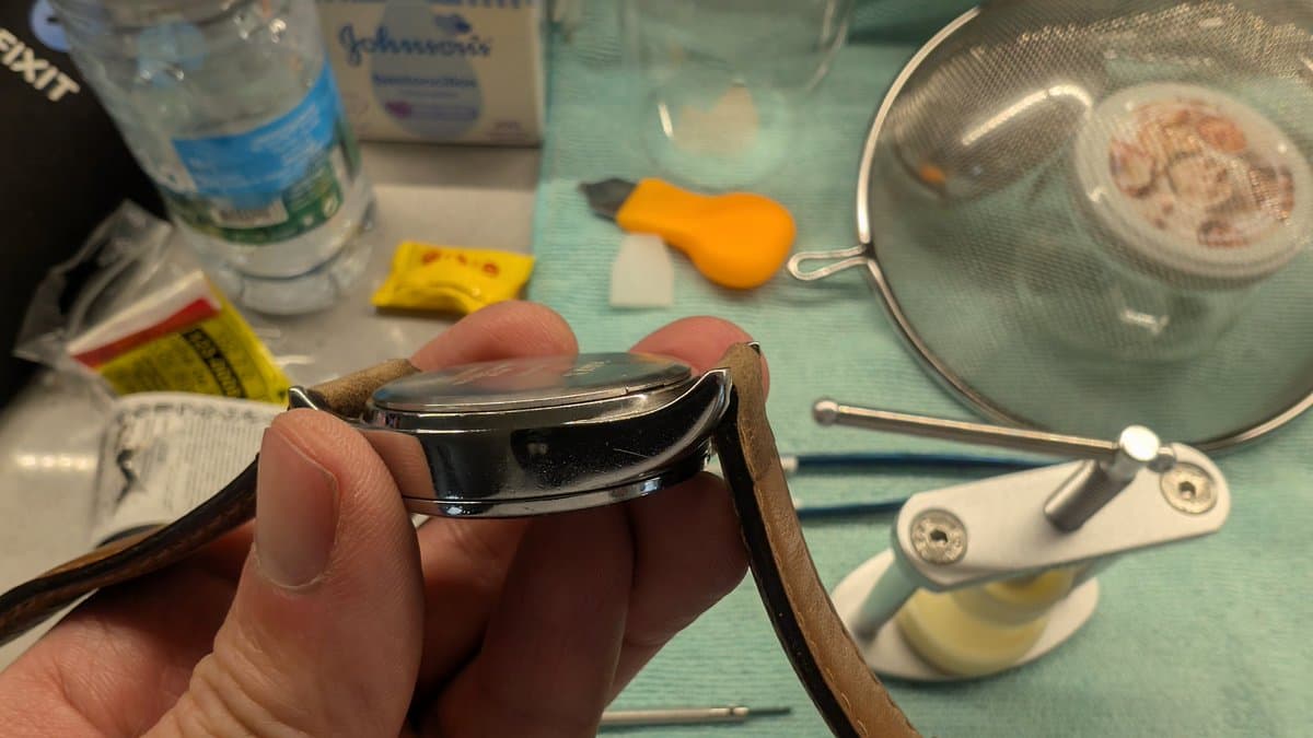 Hands holding open watch case back with snap closure tool, workbench with battery, tweezers, sieve, and Johnson's product visible.