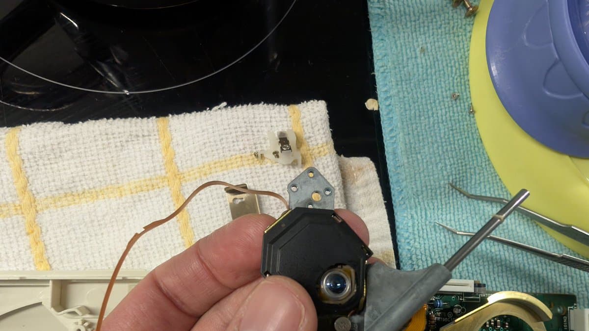 Technician holding PC Engine CD-ROM2 laser pickup assembly during disassembly, with circuit board and metal bracket visible on work surface.