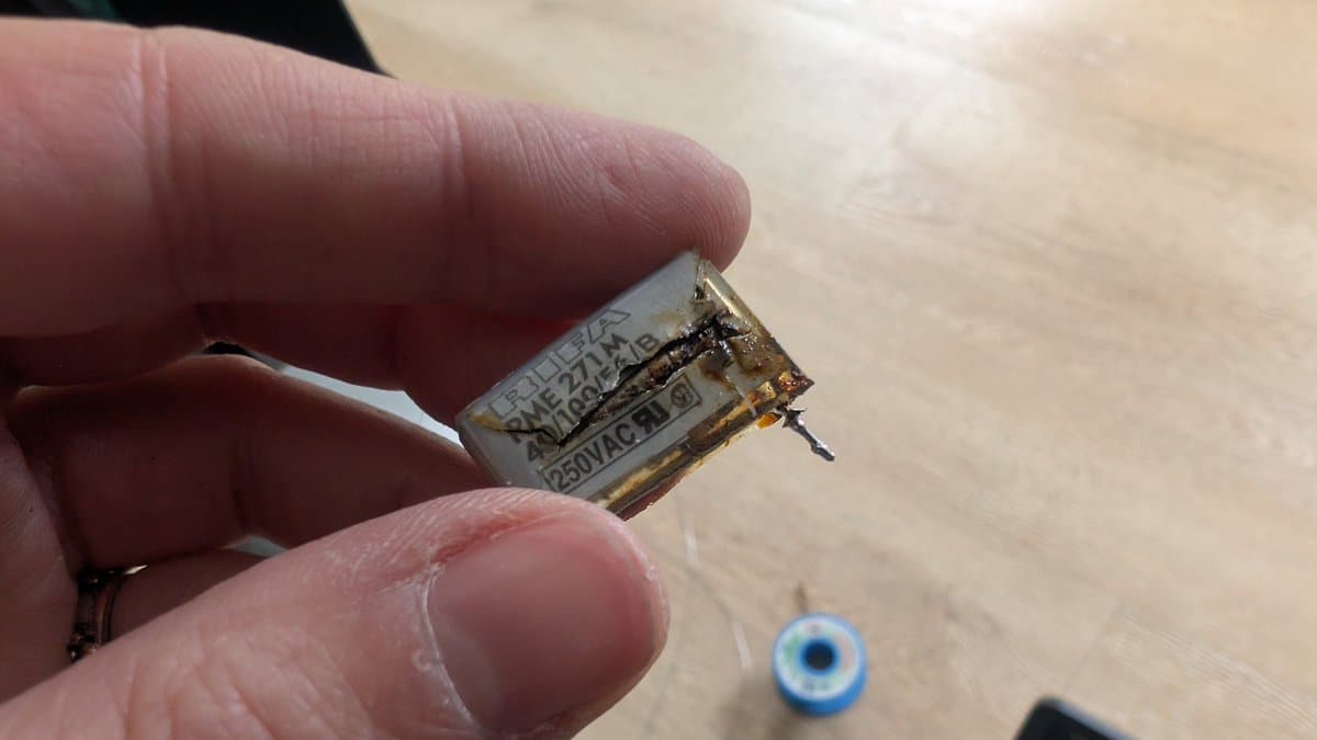 Burnt X2 safety capacitor held in fingers, showing catastrophic thermal damage and charring from the Beovision 1 repair.